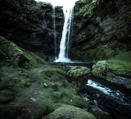 A wild natural fresh sparkling waterfall in a lonely green valley with moos covered rocks and ice cold mountain water flowing down black rocky walls on a rainy moody day in Iceland without tourists