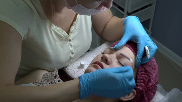 A woman-cosmetologist smears the cream of the eyebrows of a young girl after a microblading procedure in a beauty salon. Permanent makeup eyebrows. Close up. The view from the top. 4K, 25 fps.