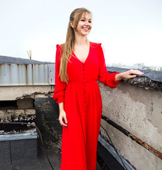 young blonde sexy girl in a red dress with long windy hair is standing and smiling  on the roof...