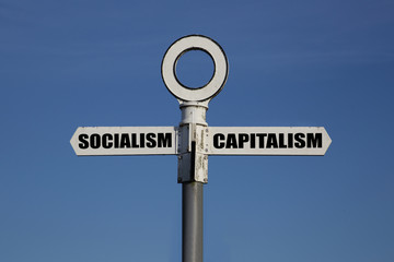 Old road sign with socialism and capitalism pointing in opposite directions against a blue sky