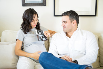 Pregnant Woman Holding Ultrasound Scan By Man On Sofa