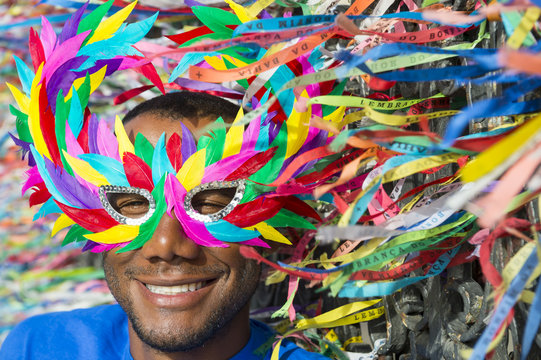 Carnival scene features smiling Brazilian man in colorful mask with wish ribbons.