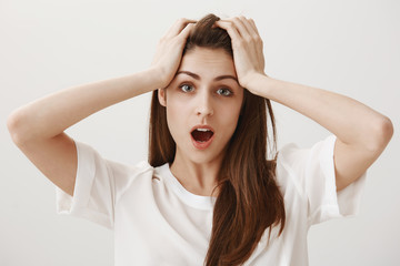 Deadline is near. Portrait of trobled and stressed attractive young woman holding hands on head and dropping jaw, being shocked with loads of work that need to tackle alone, standing over gray wall