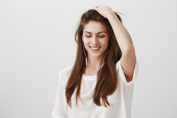 Music that recalls good memories. Portrait of attractive playful woman in white shirt touching hair and looking down with broad smile while listening music in headphones, standing over gray wall