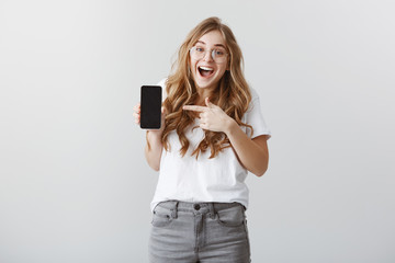 Can you imagine, finally bought new device. Satisfied excited young european girl in glasses, dropping jaw from amazement, showing black smartphone and pointing at gadget with index finger