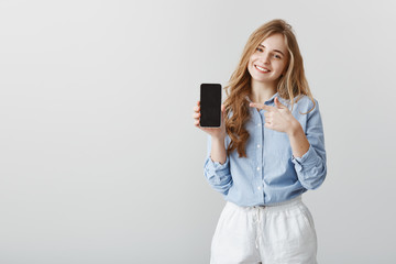 Naklejka premium Super useful device. Studio shot of pleased good-looking female student with blond hair in blue-collar shirt, showing black smarpthone and pointing at gadget with index finger, offering to buy item