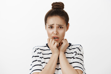 Woman scared to answer for own mistakes. Cute caucasian female brunette with bun hairstyle, frowning, biting fingers, looking anxious over white wall, feeling regret or being scared over white wall