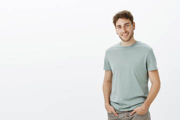Portrait of satisfied carefreee attractive male model in casual outfit, holding hands in pockets and smiling with charming expression, tilting head, being happy and in good mood over gray wall