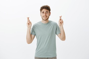 Studio shot of cute blond european male student in t-shirt raising crossed fingers and smiling with shy and awkward expression, making wish or hoping for dream come true over gray wall