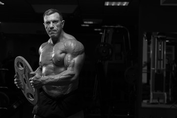 Fototapeta na wymiar High level bodybuilder with perfect relief body posing with heavy weights in hands. Strong mature man in the gym. Black and white image