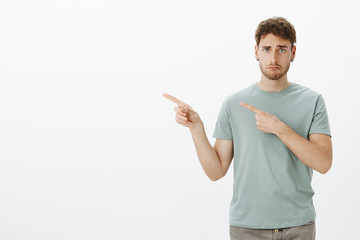 Sad gloomy young man in t-shirt, making upset disappointed face while pointing left with index fingers, frowning, being unhappy and miserable, missing chance to visit awesome place while on vacation