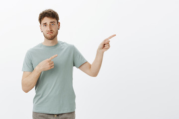 Indifferent unimpressed handsome man with bristle in earrings, pointing right with index fingers and smirking, being careless and unimpressed while discussing something boring over gray background