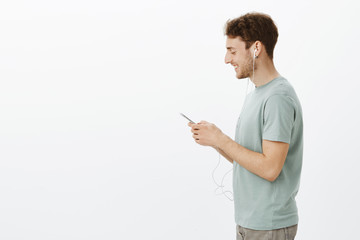 Profile shot of handsome joyful european male coworker with fair hair, holding smartphone and listening music in earphones, smiling at screen while being pleased with cute message from girlfriend