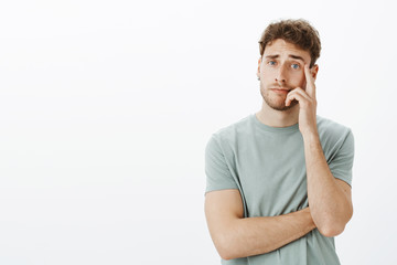 Unimpressed careless attractive young male with short fair hair, holding hand on cheek and staring with no care at camera, hearing stupid ideas during interview, standing uninterested over grey wall