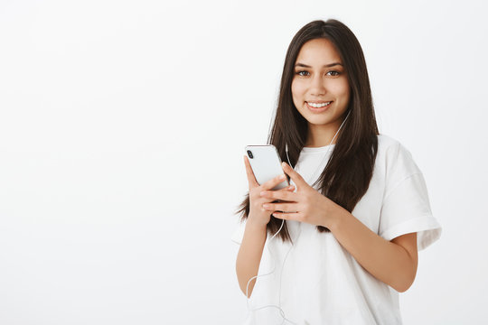 Happy beautiful Caucasian girl in white t-shirt, holding smartphone and listening music in earphones, gazing with cheerful smile at camera, picking track to go for walk, standing over gray wall