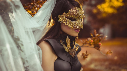 Golden, mysterious woman dressed in white with mask of leaves in copper and gold color in a garden in autumn