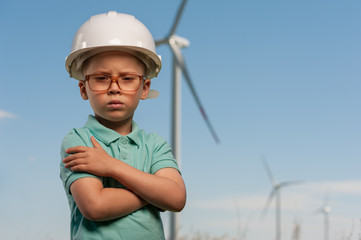 Serious smart little boy with glasses and white helmet stands against the background of windmills and the blue sky. Concept of dreams and future plans