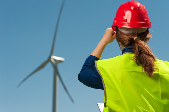 Concept of alternative energy. Rear view woman engineer in green vest and helmet looking towards windmills against the blue sky