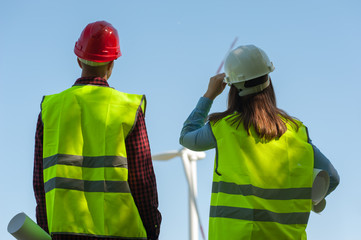 Rear-view engineers - man and woman in green vests holding helmets and looking at a windmill evaluating the work of their project