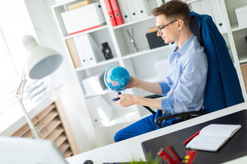 Fototapeta premium A young man sits in the office at a computer desk and holds a globe in his hands.