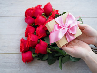 Preparing a gift, on a wooden table, with roses. Female hand holding a gift, wrapped with ribbon, on a background of red roses, the space under the text