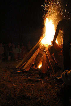 Blurred People sit at night around a bright bonfire and silhouette of a man throwing firewood into a fire