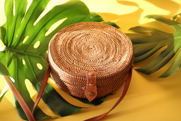 Round shaped Ata rattan boho bag with a leather strap laying on a Monstera leaf and yellow, sunny...