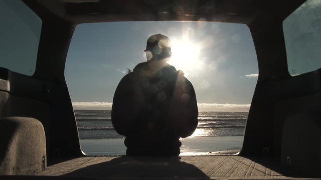 Person sitting at the back of the car watches bird fly by and ocean waves crash, parked on sandy beach in Oregon.