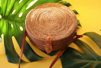 Round shaped Ata rattan boho bag with a leather strap laying on a Monstera leaf and yellow, sunny background