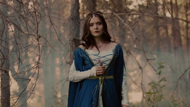 Engaging woman dressed as elvish princess walking in the forest in fog. Attractive model holding small bouquet of flowers. Marching.