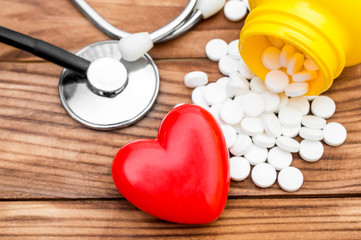 Pills scattered from plastic bottle with red heart and stethoscope on wooden background. Close up.