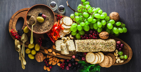 banner of Wooden serving board with vegan snacks. Pate of olives and cheese from cashew nuts with herbs. Healthy appetizer with grapes, nuts, and dried fruits on a black slate table.