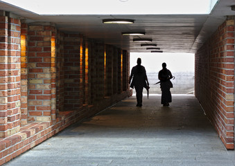 Two people silhouetted against a bright light walking through a tunnel with brick walls towards the camera