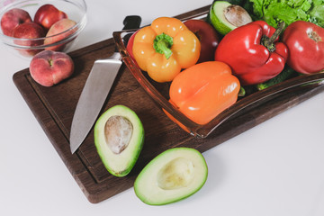 Top view of vegetables in plates prepared for slicing on a wooden board. There are sweet peppers, avocado and lettuce in a glass plate.