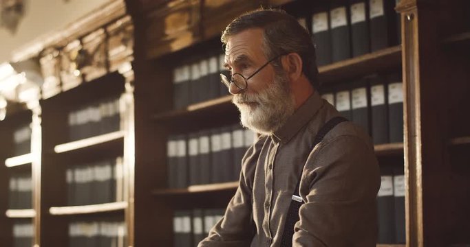 Portrait of the old Caucasian man in glasses standing in the library cabinet and smoking tabacco pipe.