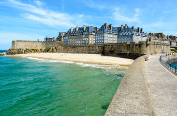 The walled city of Saint-Malo in Brittany, France, with granite residential buildings sticking out above the rampart and people sunbathing on the Mole beach at the foot of the high wall.
