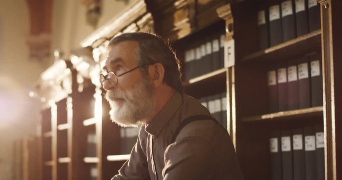 Close up of the old gray haired professor in glasses smoking tabacco pipe while standing in the library passage and looking around.