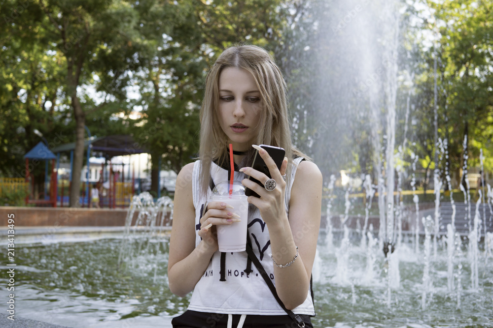 Wall mural Girl with mobile phone and cocktail in the park in front of the fountain