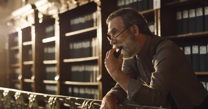 Portrait of the old gray haired man in glasses smoking tabacco pipe while standing in the library passage and thinking.