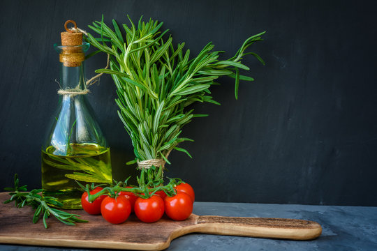 Fresh bunch of rosemary, cherry tomatoes, bottle of rosemary oil or olive oil on cutting board on stone background with copy space.