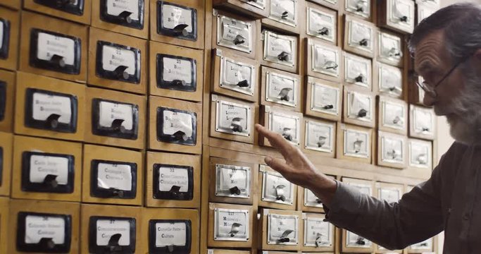 Senior man in glasses and with gray beard opening a drawer with card indexes in the library archieve.