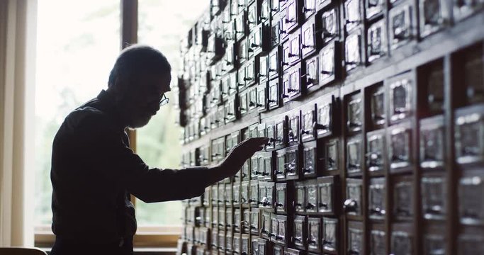 Senior man in glasses and with gray beard looking for a drawer and then opening it inside with card indexes in the library archieve in the evening.
