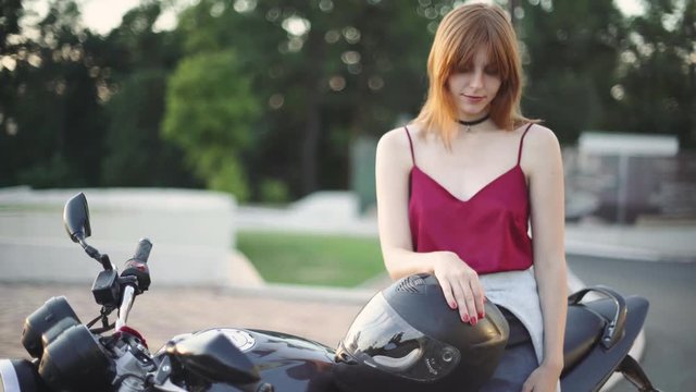 Beautiful young red-haired woman motorcyclist with her motorcycle and helmet. On street at sunset or sunrise. Woman biker 
