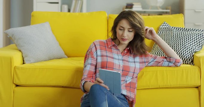 Caucasian young beautiful woman sitting on the floor near the yellow sofa and reading or watching something on the tablet computer.