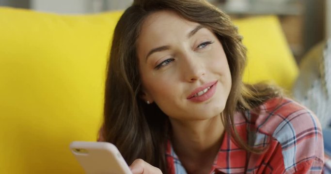 Close up of the young pretty woman lying on the couch and typing while chatting on the smartphone with a content face.