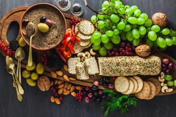 Wooden serving board with vegan snacks. Pate of olives and cheese from cashew nuts with herbs. Healthy appetizer with grapes, nuts, and dried fruits on a black slate table.