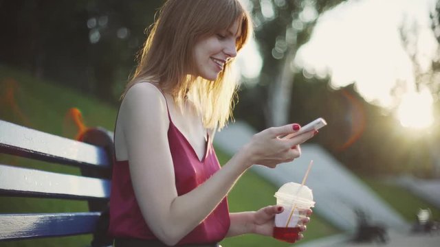Beautiful young red-haired  woman sitting on a bench in the park drinking juice and using a smartphone on sunset 