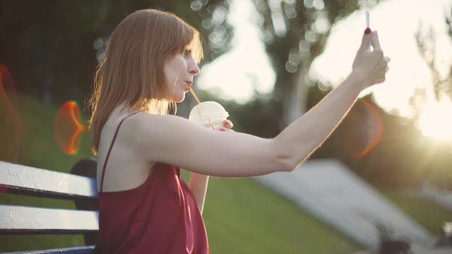 Beautiful young red-haired  woman sitting on a bench in the park drinking juice and using a smartphone on sunset 