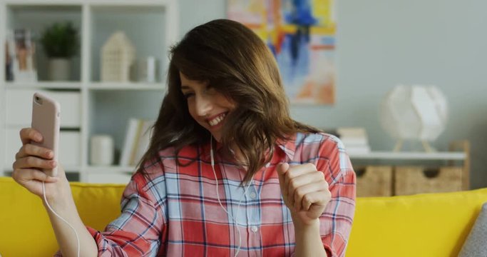 Close up of the young cheerful woman in motley shirt and headphones listening to the music on the smartphone anf moving into the rhythm.