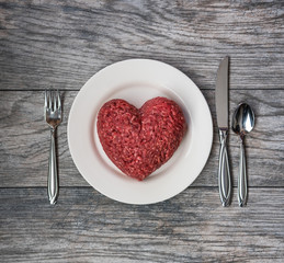Raw ground beef in the shape of a heart on a white plate with cutlery on a grey wood background. 